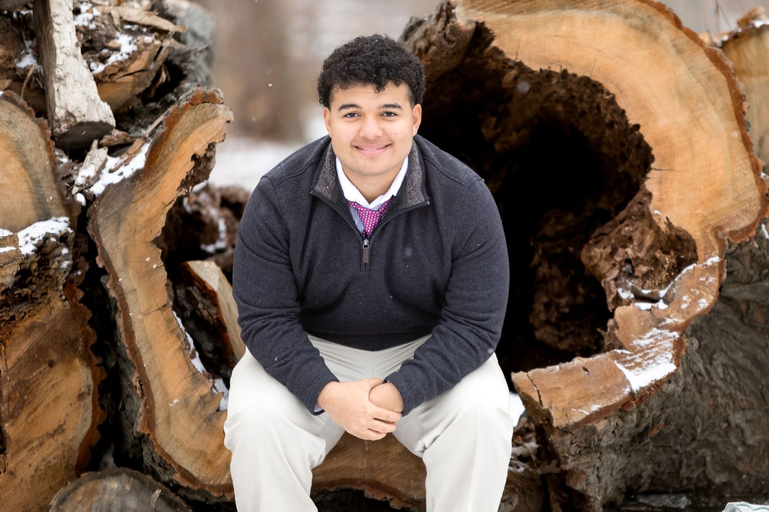A person sits on a stack of cut logs wearing a dark sweater and white pants in a casual outdoor pose.