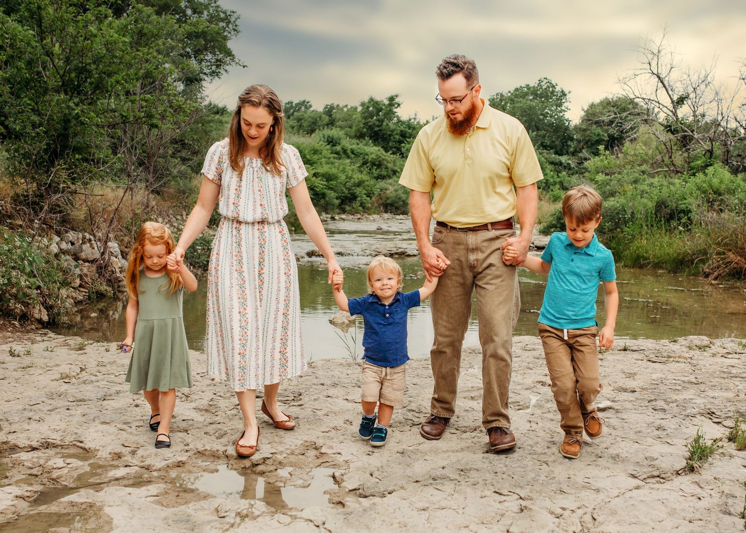 A family of five walking together and holding hands on a rocky riverbank surrounded by green trees in Fort Worth, TX.