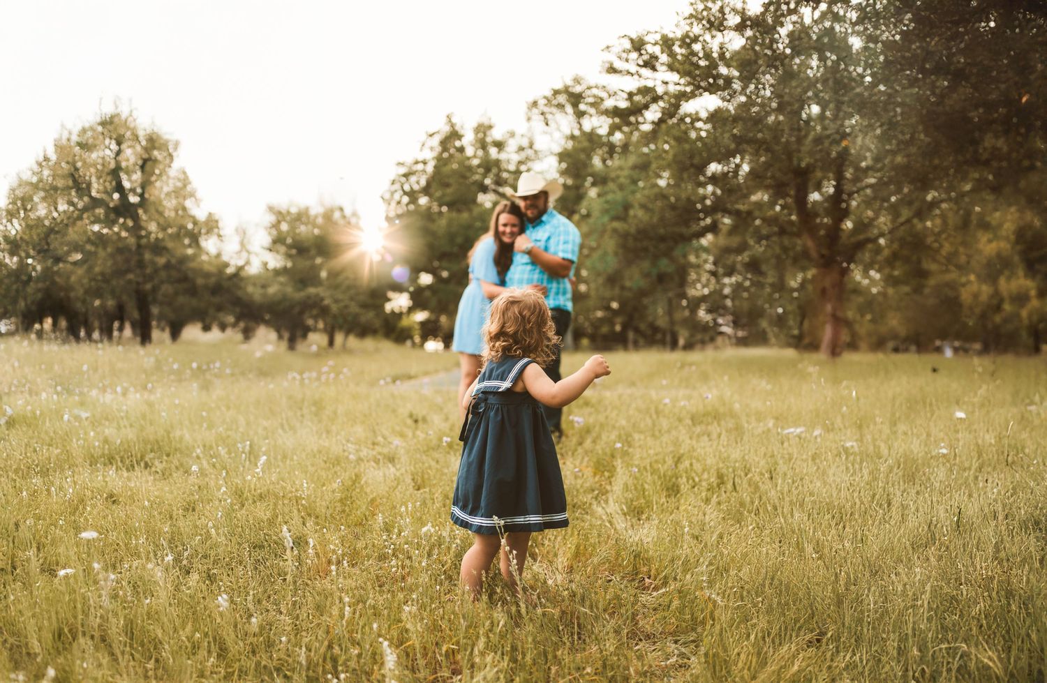 Young girl in navy dress walks toward parents in sunlit meadow with golden bokeh bubbles.