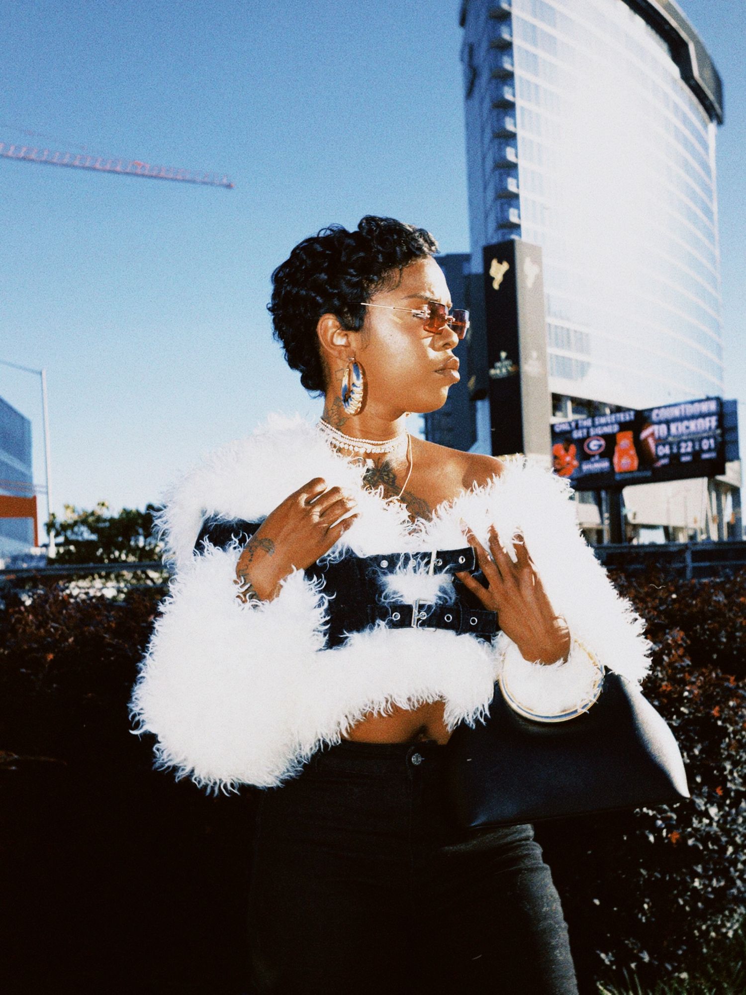 A person in a black outfit with white feather boa poses dramatically against an urban backdrop with skyscrapers.