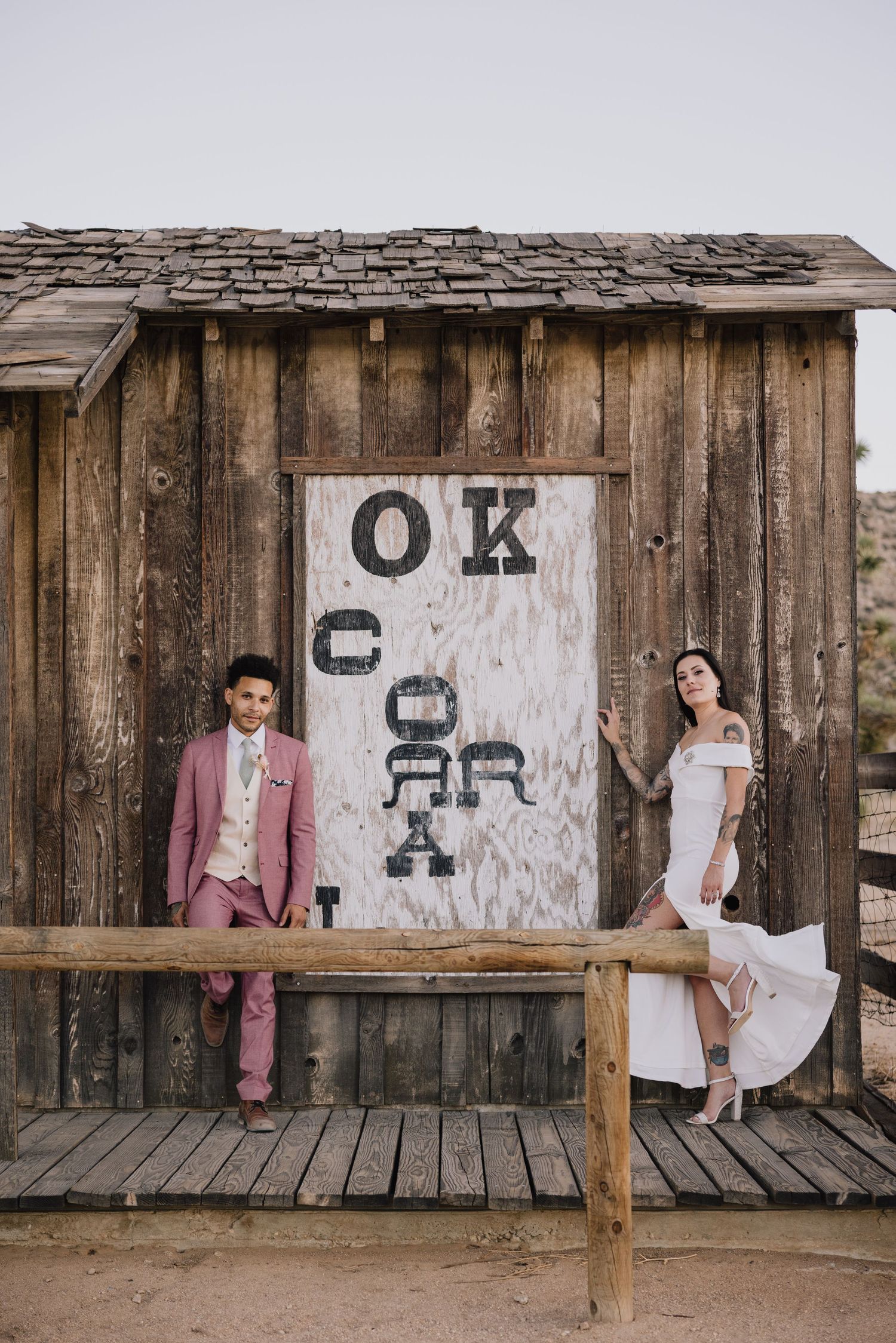 Bride and groom posing at rustic OK Corral wooden building in desert setting.