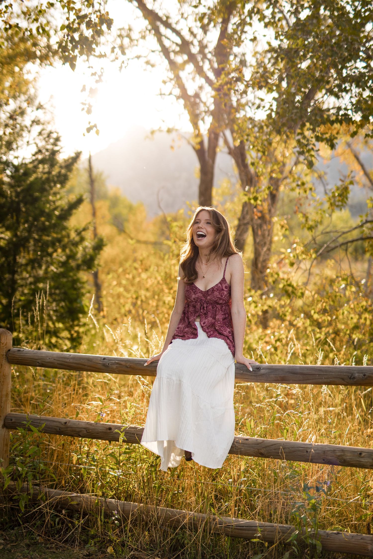 A person in a floral dress sits on a rustic wooden fence at sunset with golden light filtering through autumn trees.