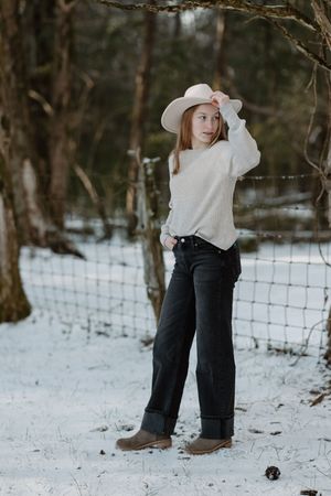 A young girl in a cream sweater and black jeans adjusts her white hat in snowy woods.