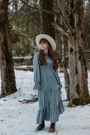 A woman in a teal maxi dress and wide-brim hat poses in a snowy forest setting.