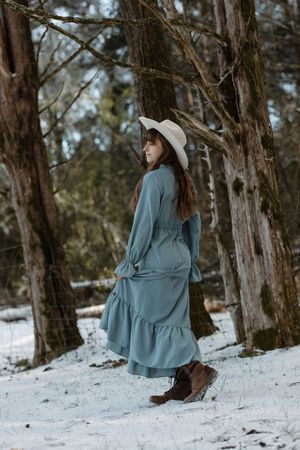 A woman in a teal maxi dress and white hat walks through a snowy winter forest.
