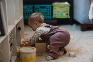 Curious toddler in purple overalls crouching to play with wooden toys on carpet.
