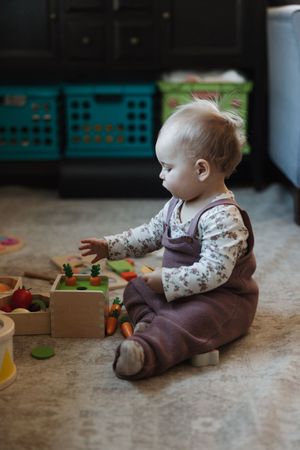 Baby in brown overalls sitting on floor playing with colorful wooden vegetable toys.