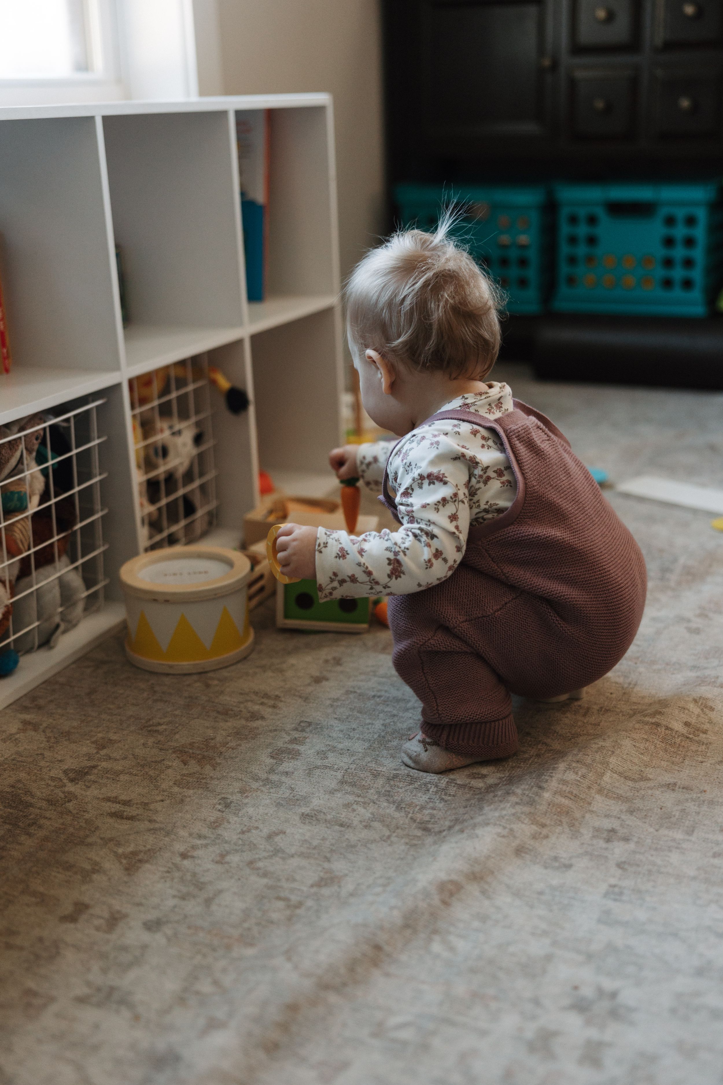 Toddler in brown overalls playing with wooden toys near white shelf organizer.