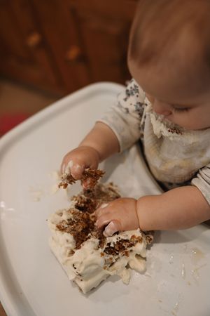 Baby in floral bib smashing first birthday cake on white high chair tray.
