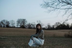 Young woman in green top and white jeans crouching in open field at dusk.