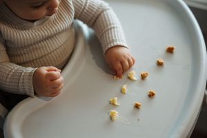 Baby in high chair exploring small food pieces on white tray during mealtime.