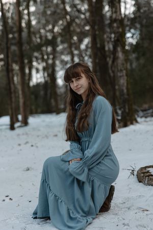 A young woman in a blue maxi dress sits on a log smiling in a snowy forest.