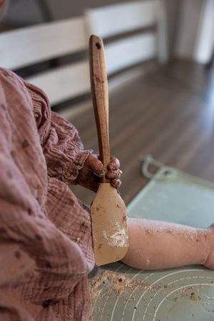 A baby in a pink outfit holds a wooden spatula covered in batter on a green mat.