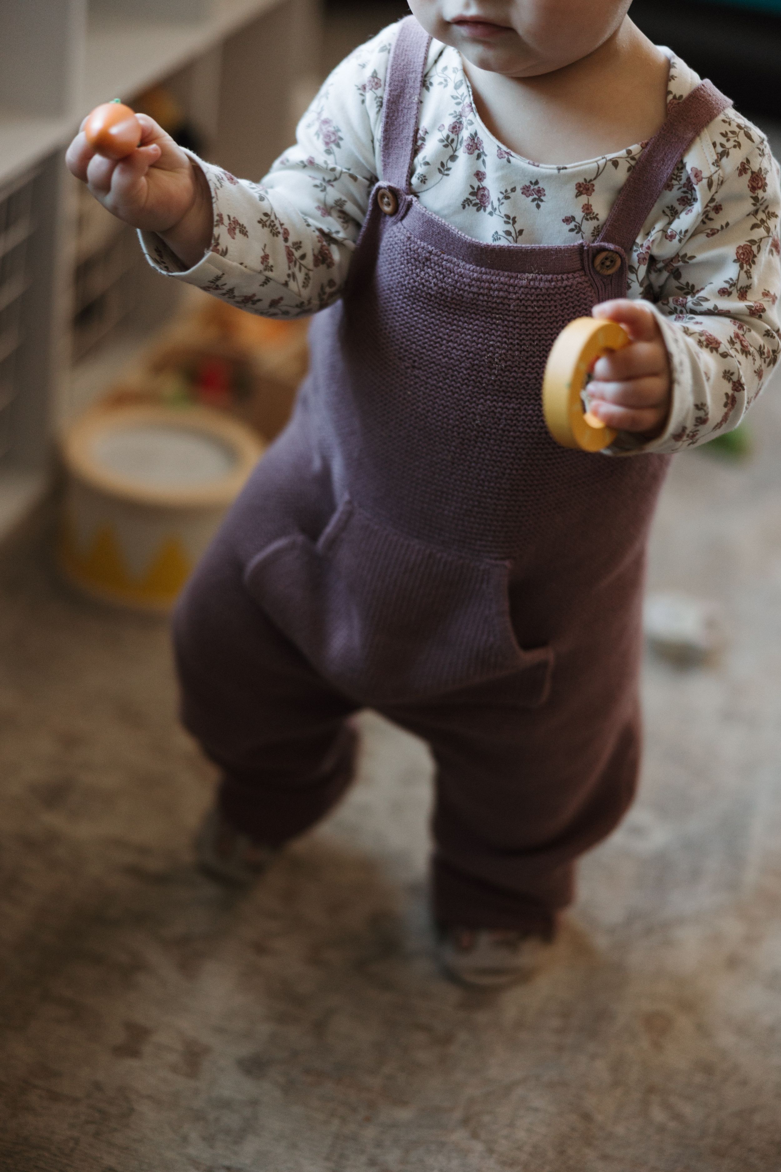 Baby in floral onesie and brown overalls taking first steps while holding toy.