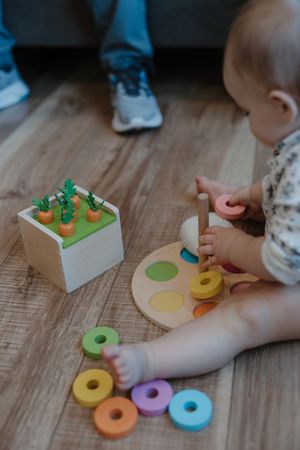 Baby plays with colorful wooden stacking ring toy and carrot puzzle on hardwood floor.