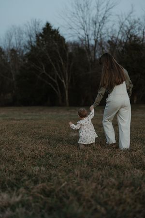 Mother and toddler holding hands walking through grassy field at twilight.