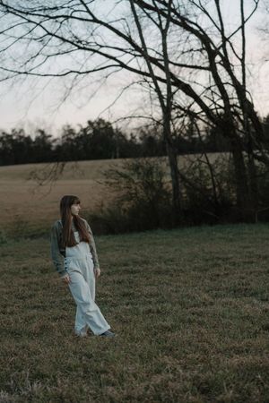 Young woman in green jacket and white pants standing near bare trees in field.