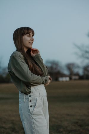 Young smiling woman in green jacket and white pants standing in open field at dusk.