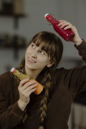 Smiling young woman with braids holds two colorful juice bottles in kitchen.