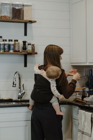 Mother carries baby in arms while cooking in modern white kitchen.