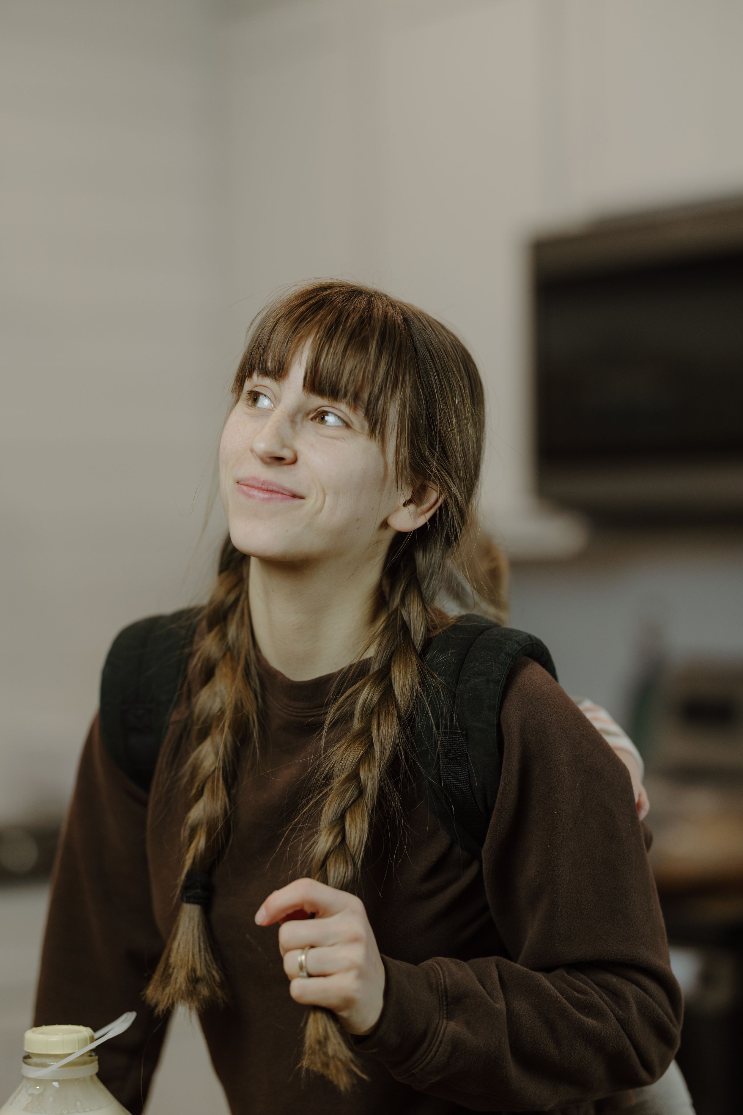 Young woman with braids in brown sweater smiles thoughtfully in kitchen setting.