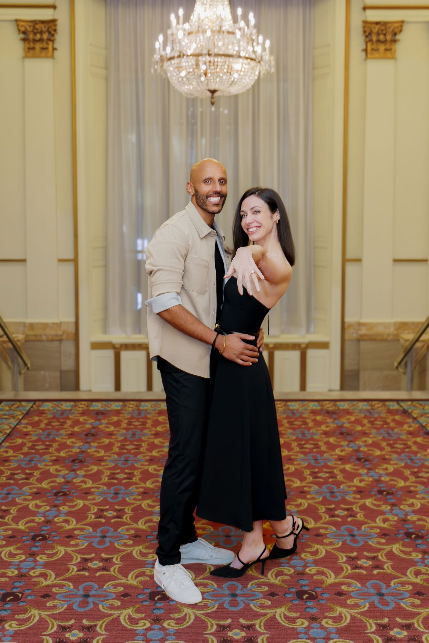 Couple dancing together in an elegant ballroom with ornate chandelier and patterned carpet.