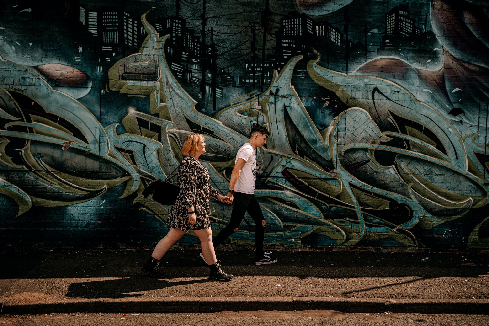 Two people walk past a large abstract graffiti mural on a dark wall casting long shadows on the ground.