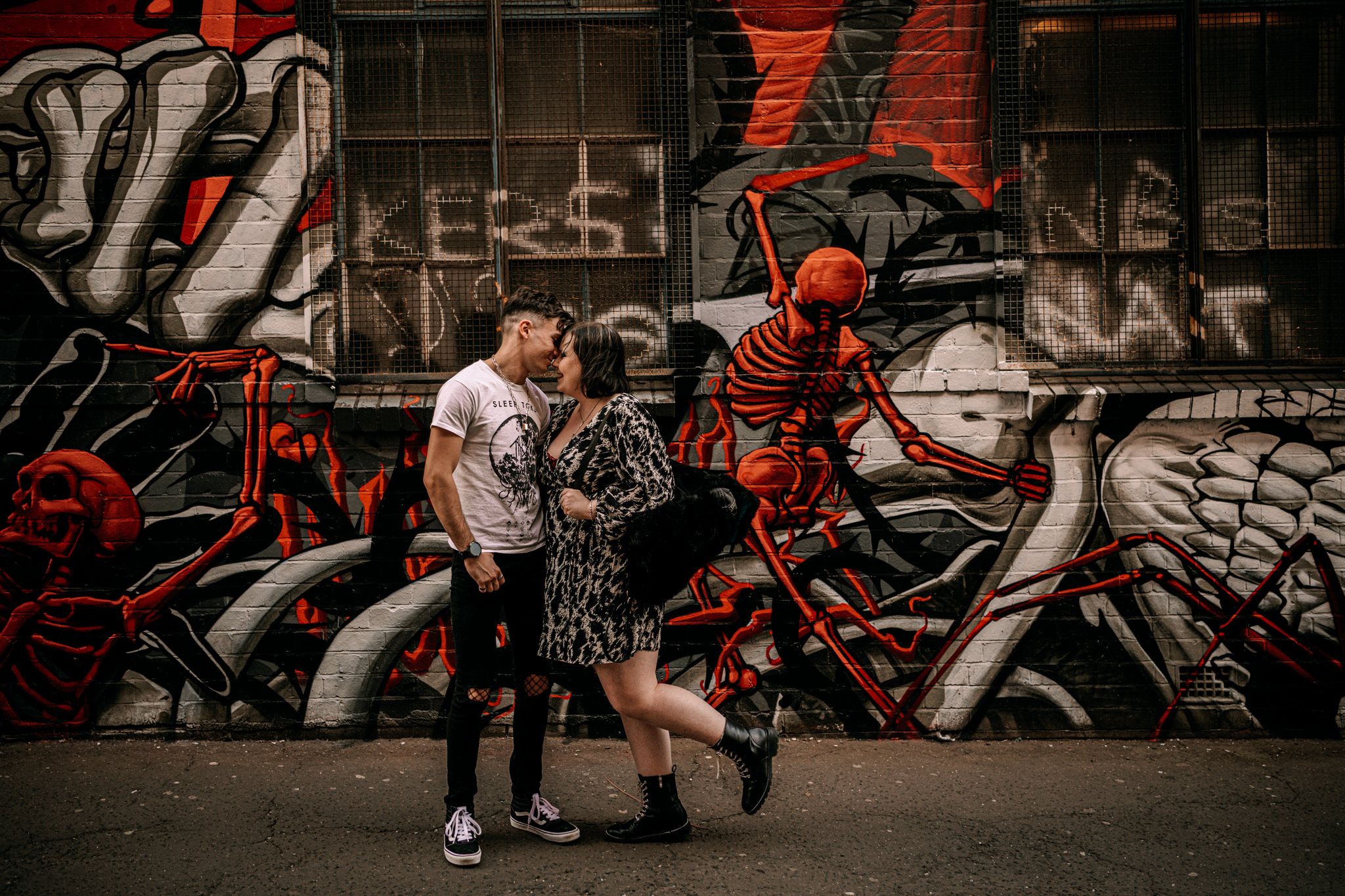 A couple poses against a vibrant graffiti wall featuring red and black street art in an urban setting.