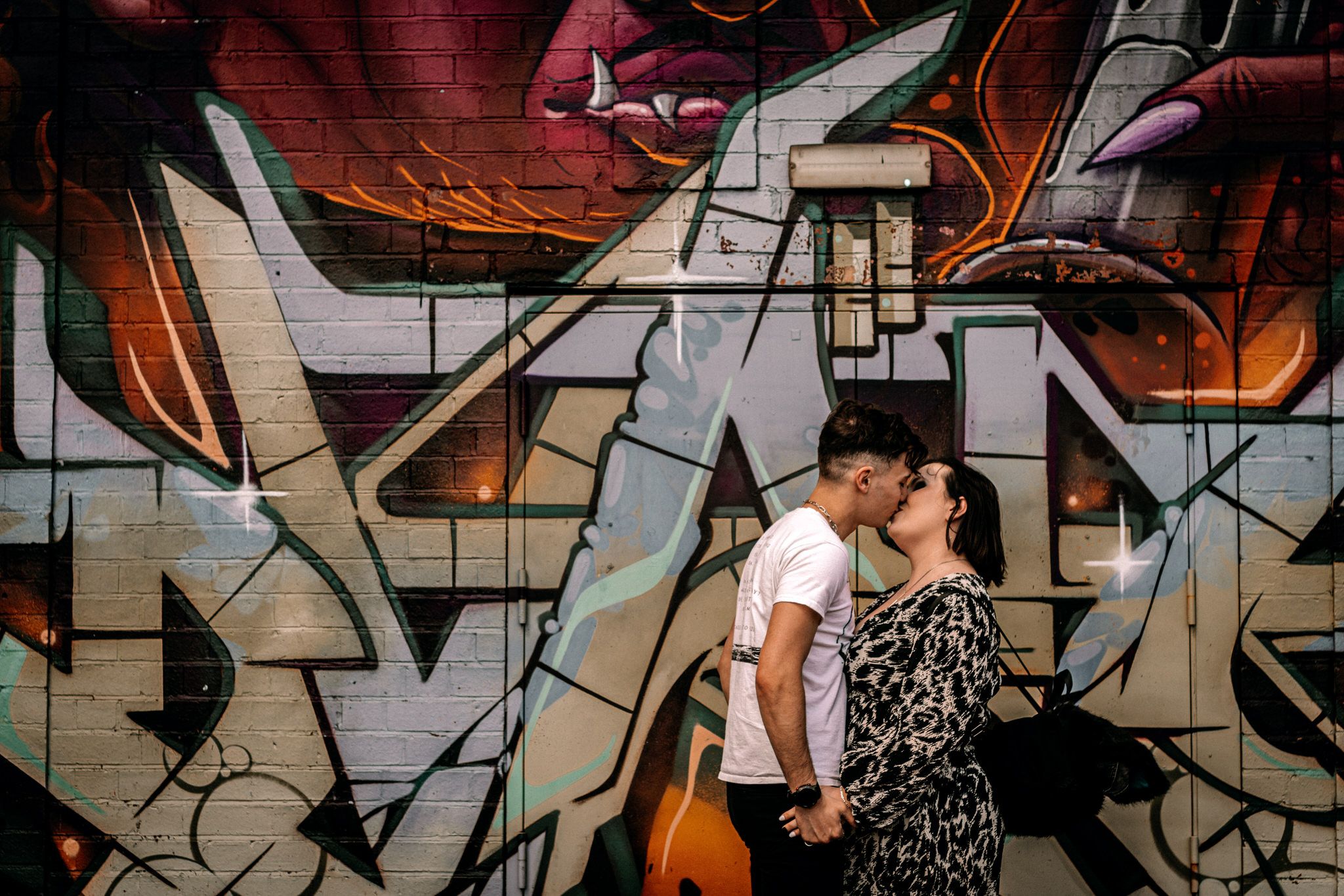 A couple poses together against a graffiti wall background in an urban setting.