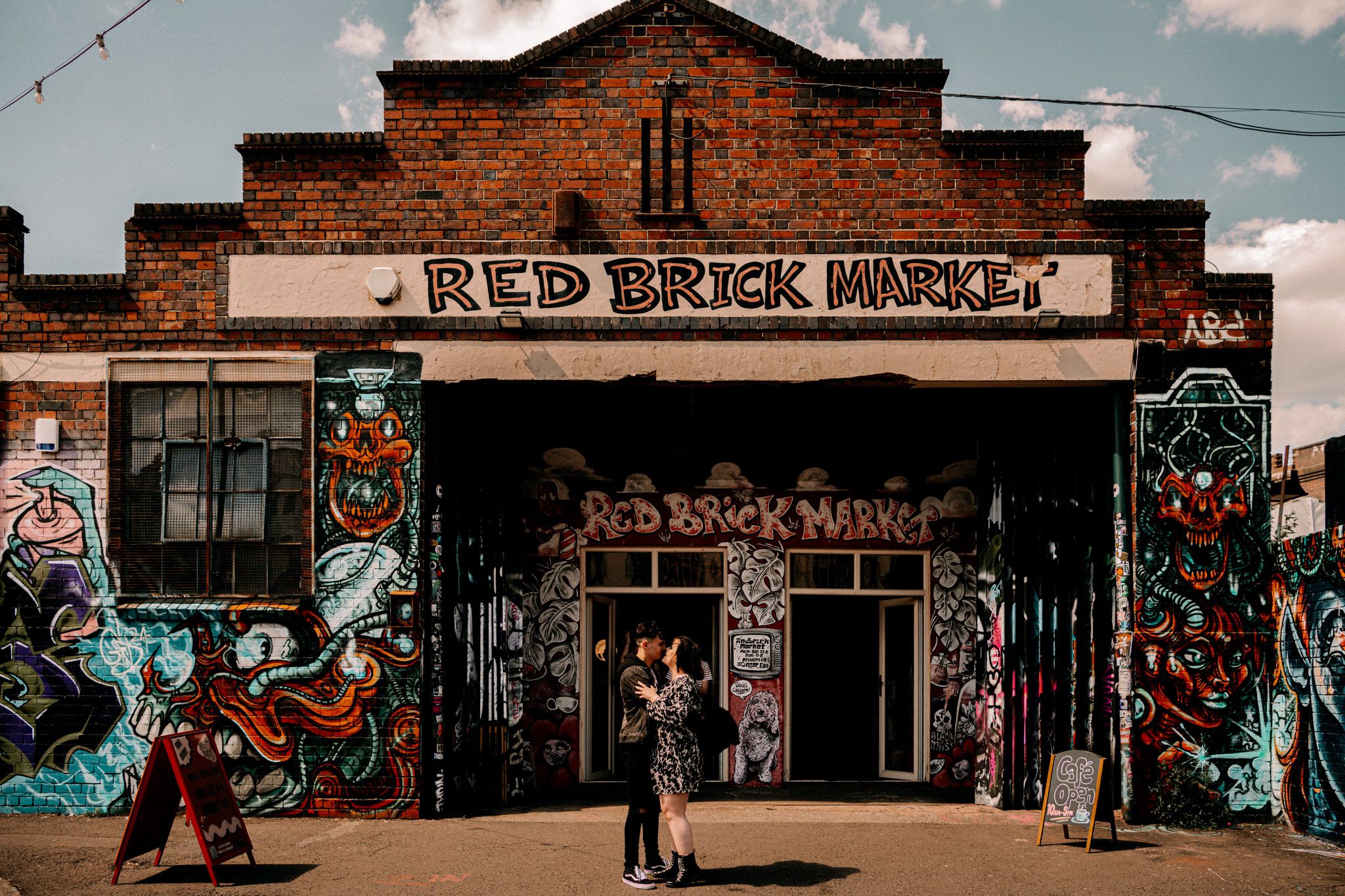 A panoramic view of a rustic red brick market building with street art murals decorating its exterior walls.