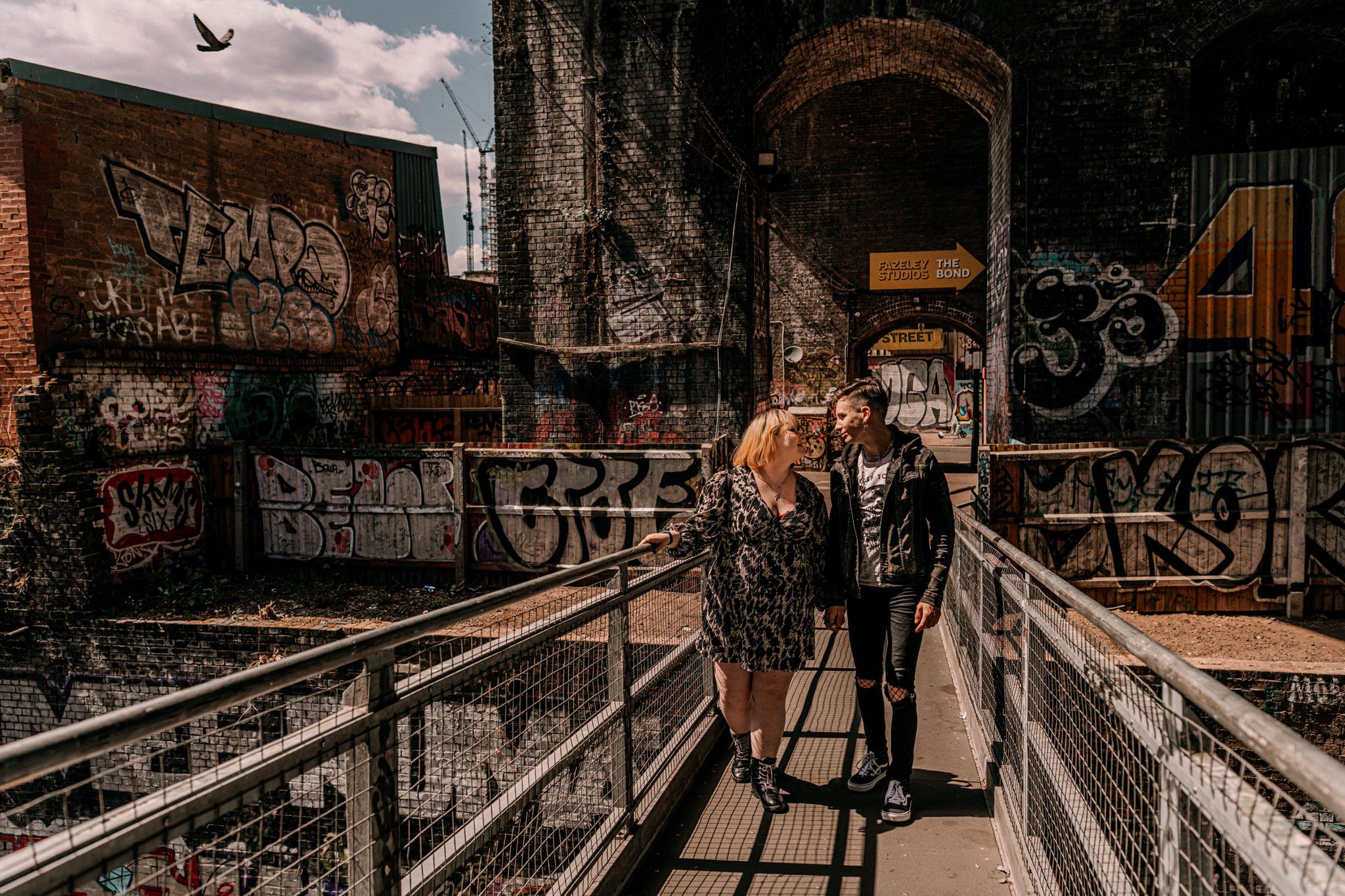 A couple walks along an industrial walkway covered in urban graffiti under a cloudy sky.