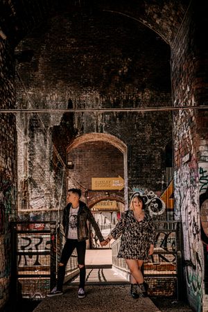 Two figures stand in a dark brick archway covered with graffiti and street art.