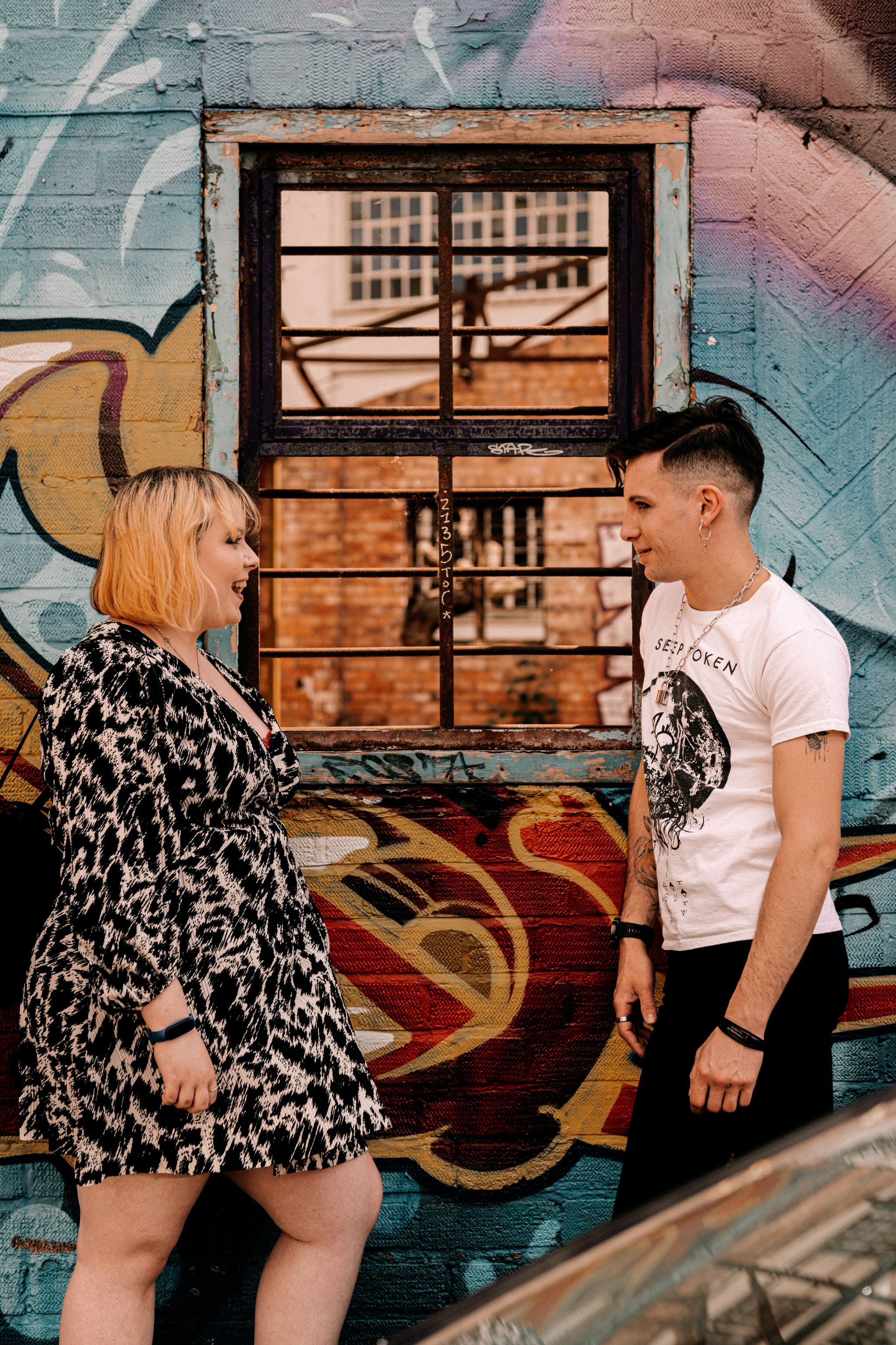 Two people pose in front of a graffiti wall with a window, creating an urban street art atmosphere.