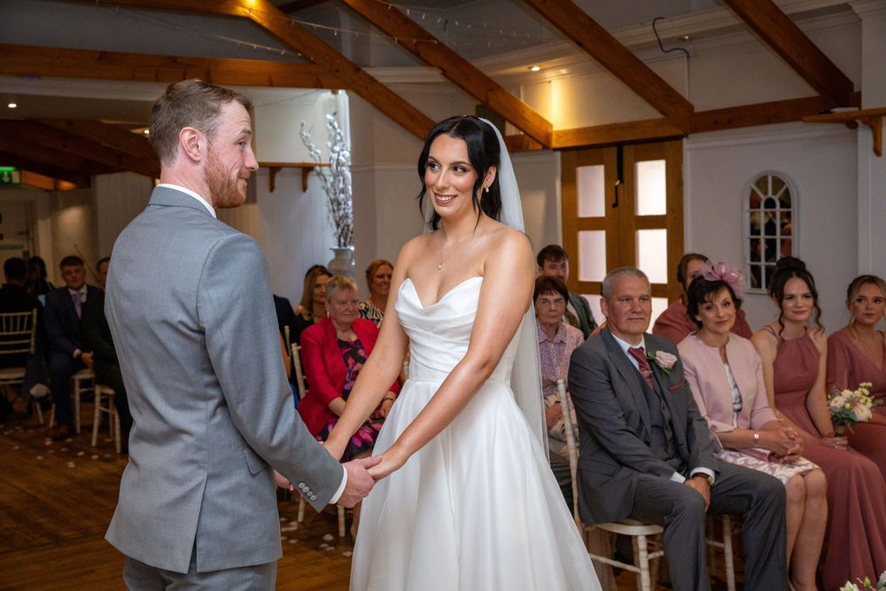 Bride and groom holding hands and smiling during intimate indoor wedding ceremony with seated guests.