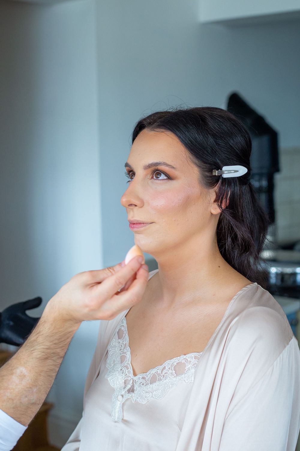 Bride having professional makeup applied while wearing white lace robe before wedding ceremony.
