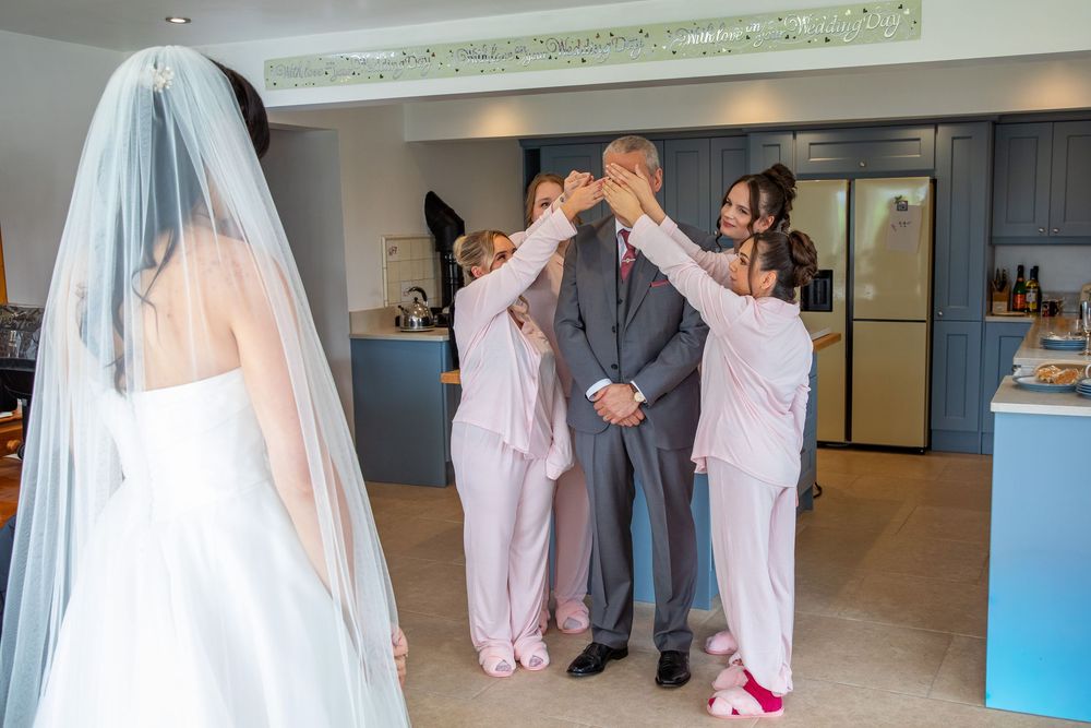 Bridesmaids in pink robes covering groom's eyes as bride approaches in elegant wedding venue.