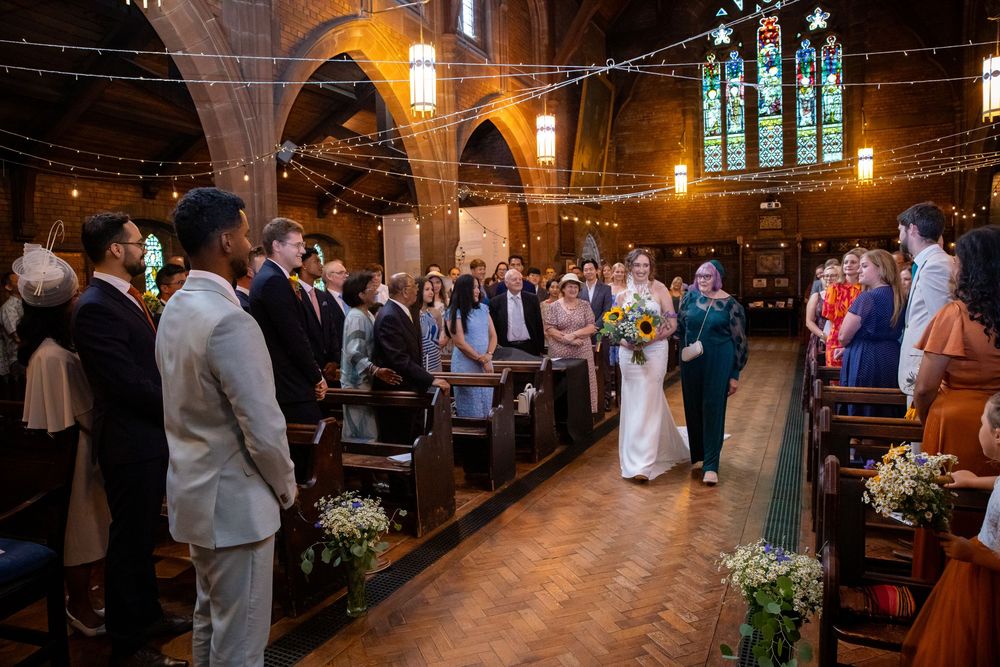 Bride walks down ornate church aisle with fairy lights and stained glass windows during ceremony.