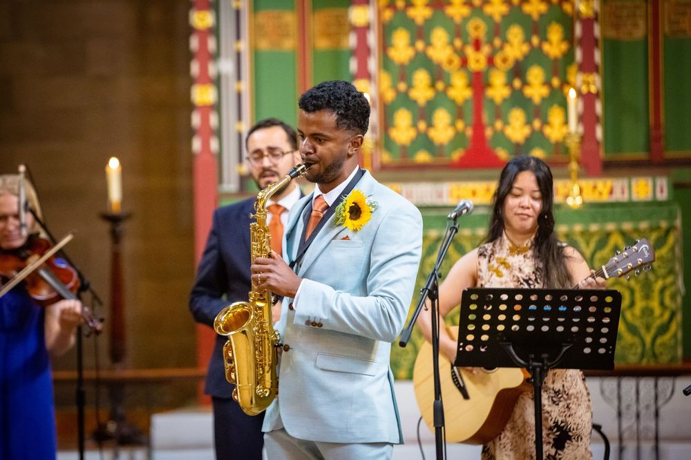 Saxophonist in light blue suit performs with band featuring violin and guitar during church wedding.