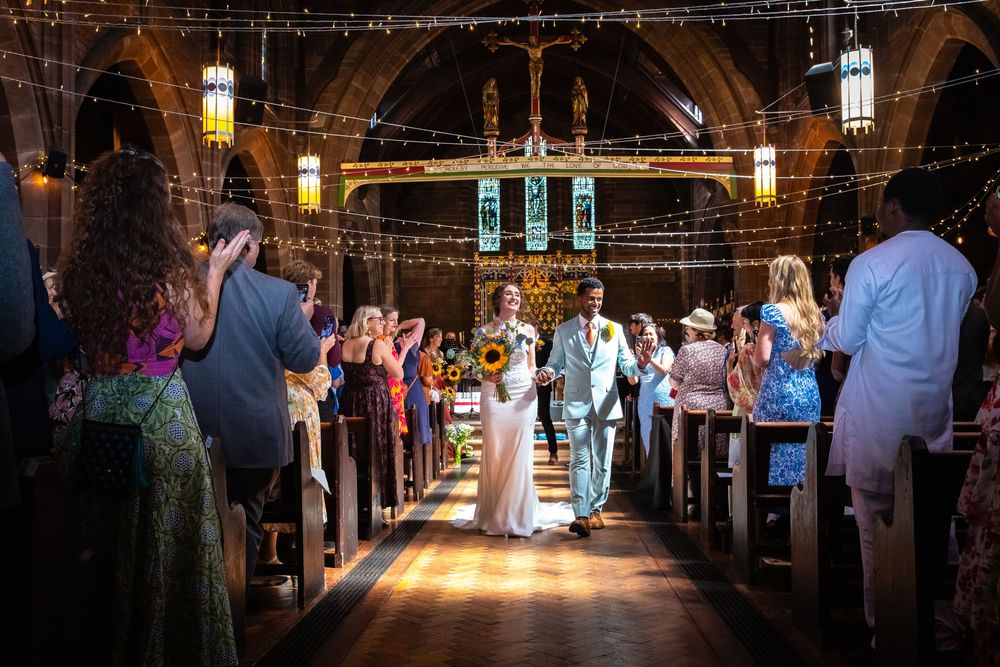Bride and groom walk down the aisle of a decorated church surrounded by cheering wedding guests.