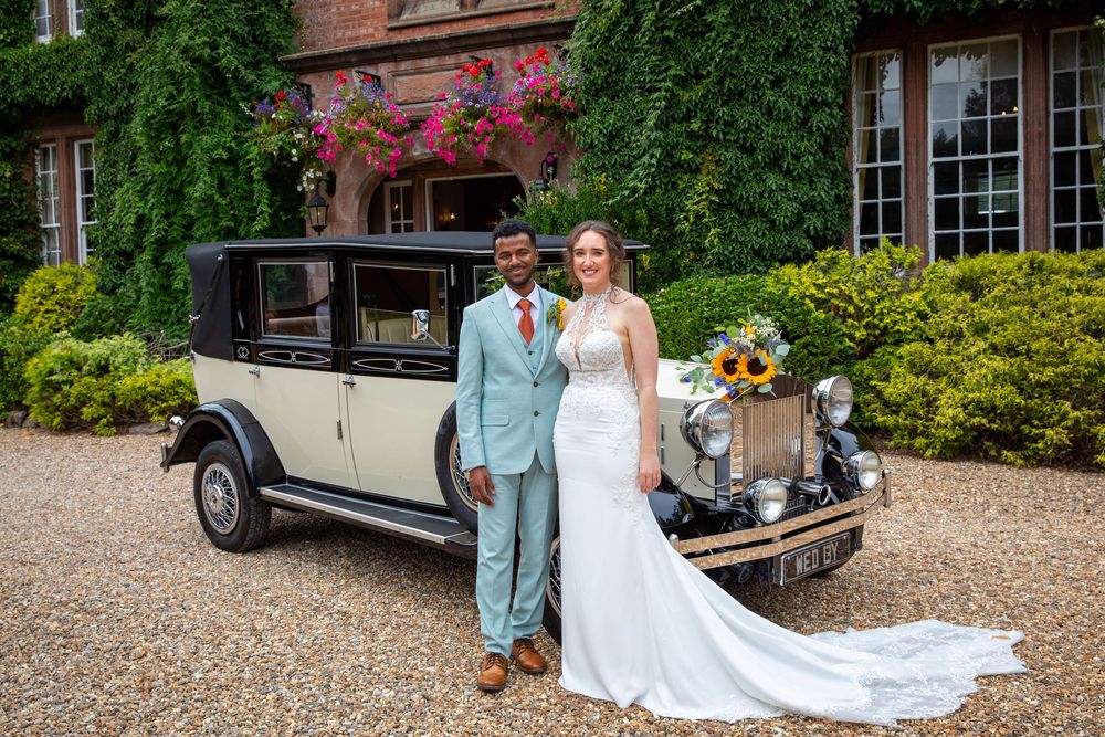 Bride and groom pose beside a vintage cream and black wedding car outside a grand manor house.