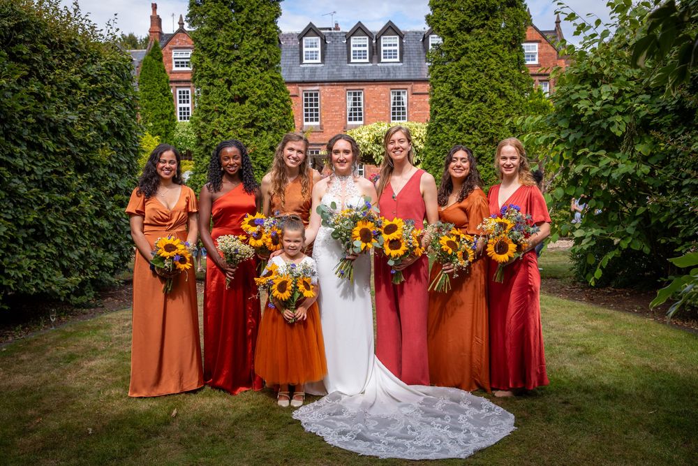 Bride stands with six bridesmaids in orange and red dresses holding sunflower bouquets outdoors.
