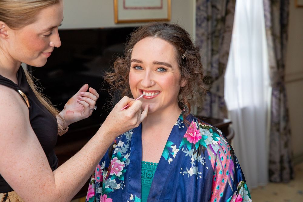 Makeup artist applies lipstick to bride wearing floral kimono robe during wedding morning preparations.