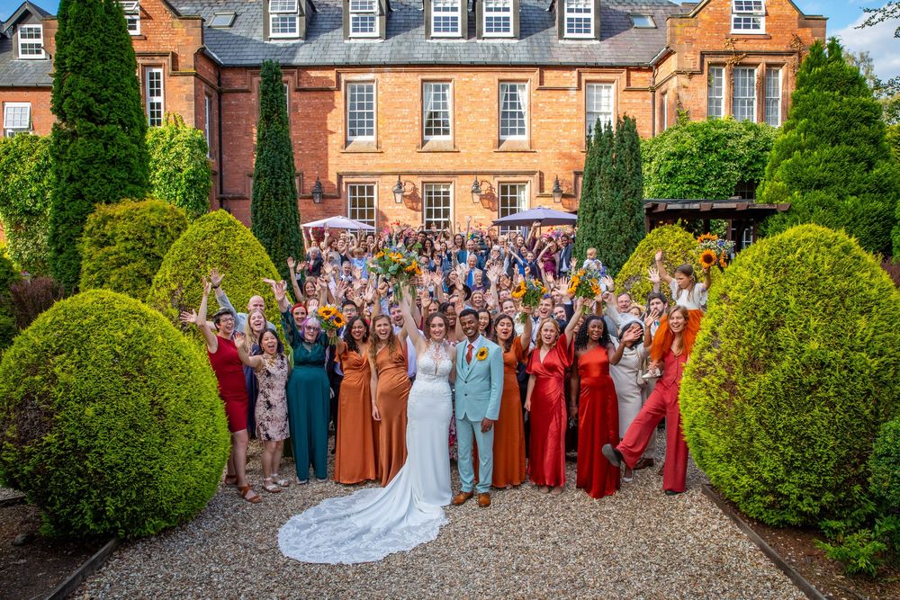 Large wedding party group photo with bride and groom in front of a red brick manor house.