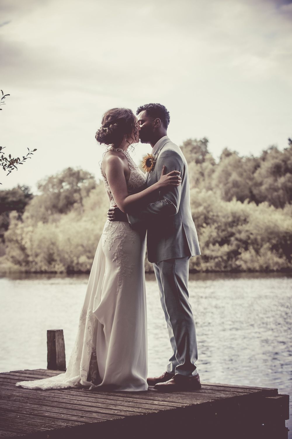 Bride and groom share a romantic kiss on a wooden dock overlooking a serene lake.