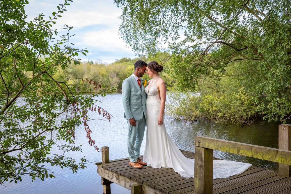 Bride and groom stand together on a wooden pier surrounded by lush greenery and a calm lake.