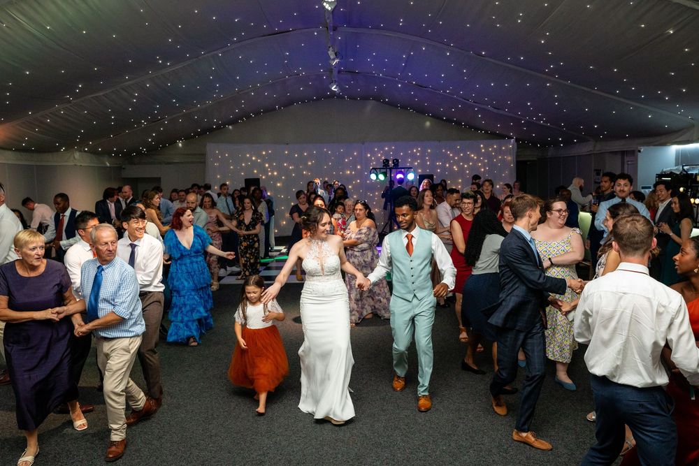 Bride, groom, and guests dance together joyfully under a starlit marquee at the wedding reception.