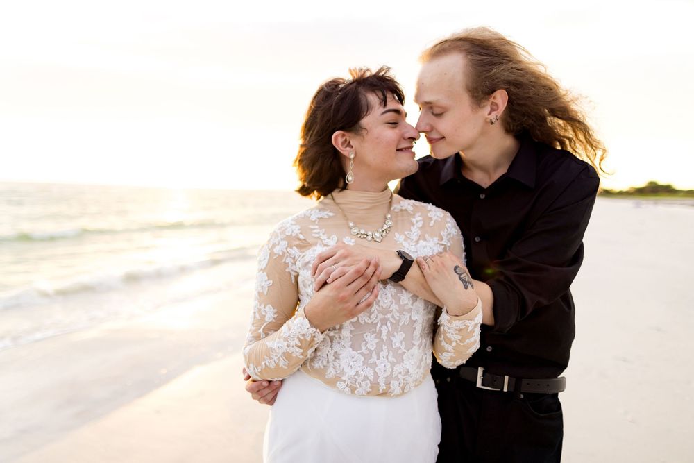 A couple shares a romantic moment in formal attire on a beach at sunset.