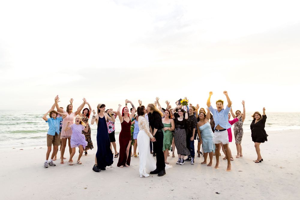 A joyful wedding party celebrates together on a white sandy beach at sunset.