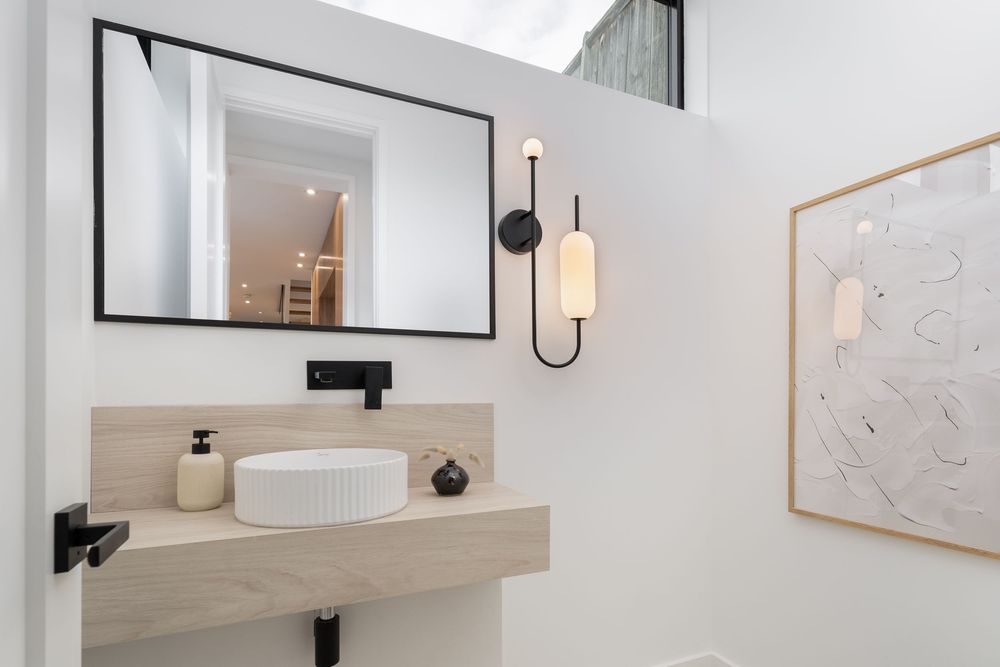 Modern white bathroom interior with minimalist design featuring a round mirror, black fixtures, and wooden shelf accents.