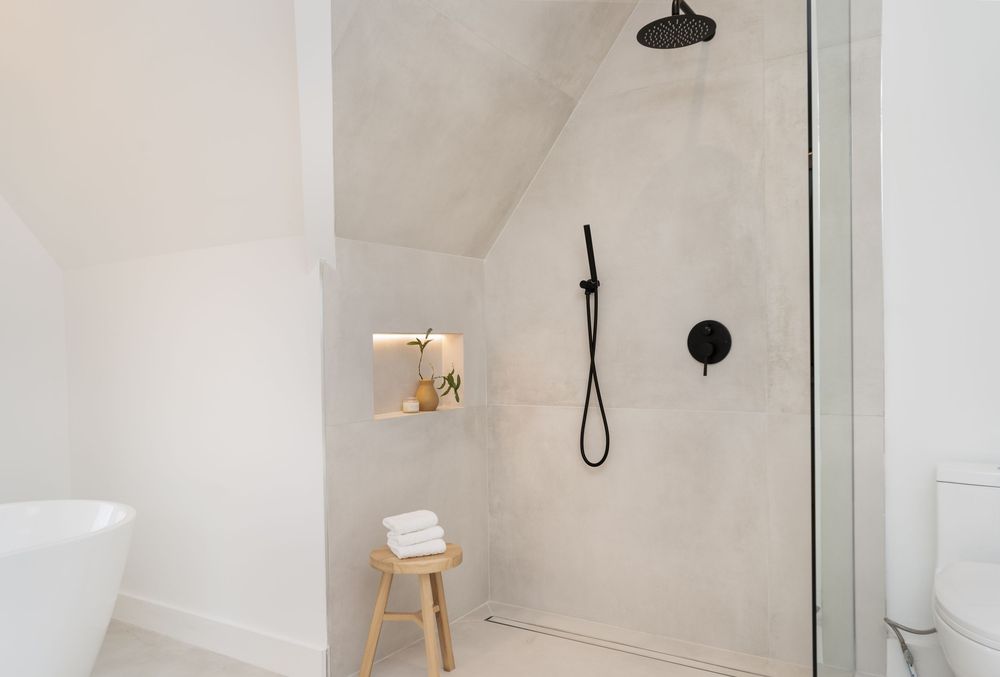 Modern bathroom interior with white walls, black rainfall shower head, and minimalist wooden stool accent.